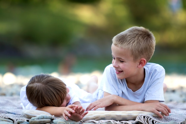 Dos jóvenes felices lindos rubios niños riendo, niño y niña, hermano y hermana que se divierten en la playa de guijarros sobre fondo de verano soleado brillante borrosa bokeh. Amistad y concepto de vacaciones perfectas.