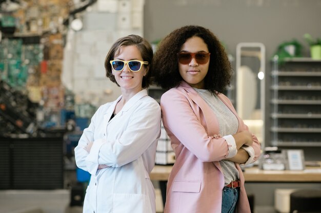 Dos jóvenes hembras multiculturales en gafas de sol de pie cerca uno del otro dentro de la tienda de óptica