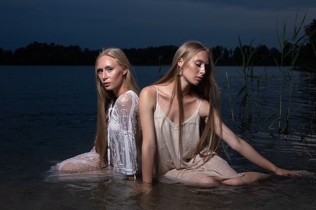 Dos jóvenes hermanas gemelas con largos cabellos rubios posando en vestidos ligeros en el agua del lago por la noche