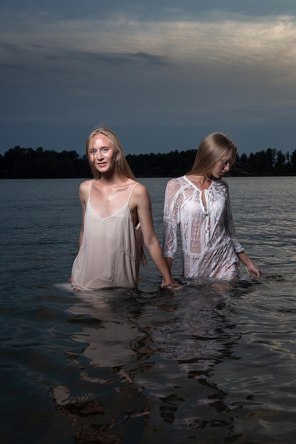 Dos jóvenes hermanas gemelas posando en vestidos ligeros en el agua del lago en la noche de verano