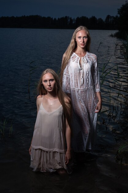 Dos jóvenes hermanas gemelas posando en vestidos ligeros en el agua del lago en la noche de verano