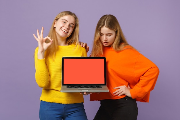 Dos jóvenes hermanas gemelas rubias chicas mostrando gesto OK, sostienen la computadora portátil con pantalla vacía en blanco aislada en la pared azul violeta pastel. Concepto de estilo de vida familiar de personas. .