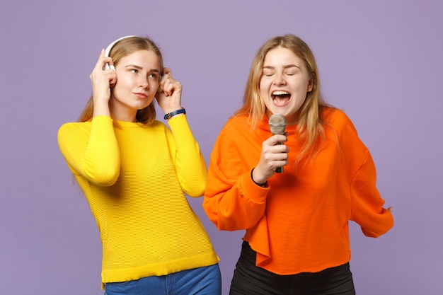 Dos jóvenes hermanas gemelas rubias niñas en ropas coloridas escuchan música con auriculares, cantan canciones en el micrófono aislado en la pared azul violeta. Concepto de estilo de vida familiar de personas.
