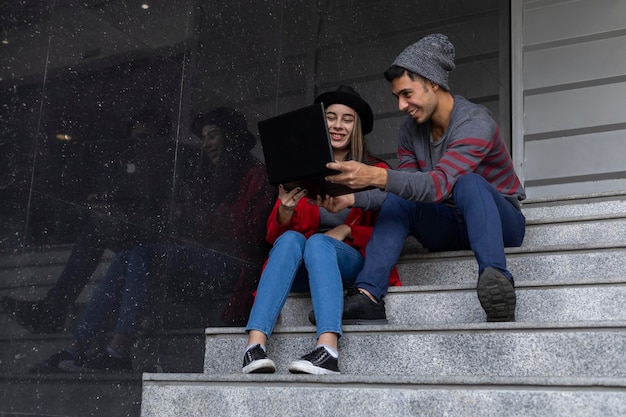 Dos jóvenes hermanos latinoamericanos sentados en las escaleras con una computadora portátil y un teléfono celular mientras comparten un tiempo de trabajo viendo videos y mensajes Concepto de tecnología