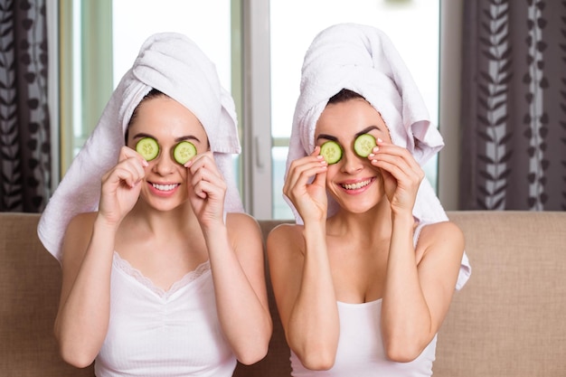 Dos jóvenes hermosas amigas sonrientes con toallas en la cabeza se aplican anillos de pepino en los ojos y hacen una máscara cosmética