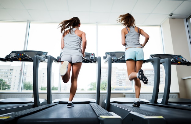 Dos jóvenes hermosas mujeres delgadas en ropa deportiva corriendo en cintas de correr en el gimnasio con ventanas y la ciudad en el fondo