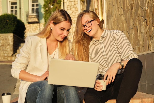 Dos jóvenes hermosas mujeres divirtiéndose mirando el monitor de la computadora portátil, mujeres de negocios sentadas en los escalones de la calle de la ciudad, estudiantes universitarios riéndose descansando