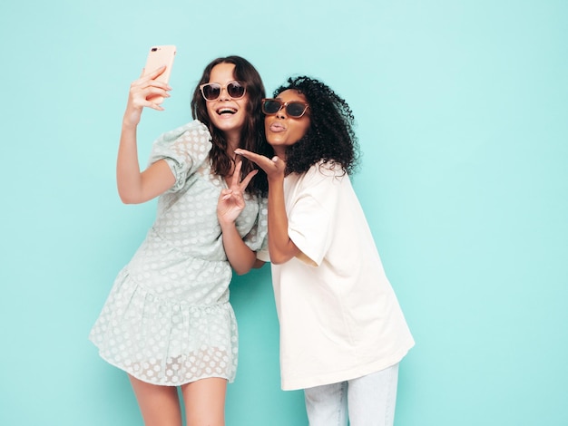 Dos jóvenes hermosas mujeres hipster internacionales sonrientes en ropa de verano de moda Mujeres sexy despreocupadas posando junto a la pared azul en el estudio Modelos positivos divirtiéndose Concepto de amistad