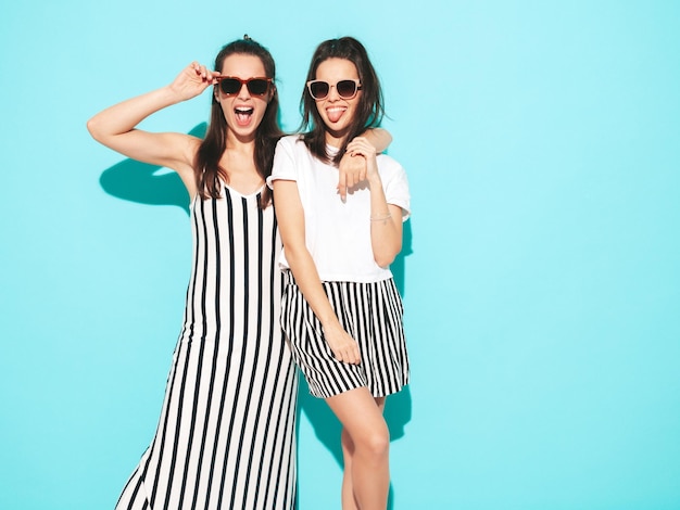 Dos jóvenes hermosas mujeres hipster rubias sonrientes en ropa de verano de moda Mujeres sexy despreocupadas posando junto a la pared azul en el estudio Modelos positivos divirtiéndose Alegres y felices con gafas de sol