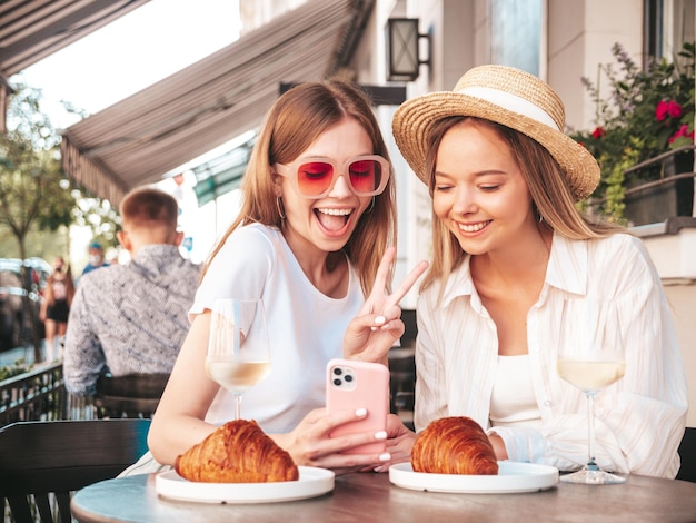 Dos jóvenes hermosas mujeres hipster sonrientes vestidas de moda de verano Mujeres despreocupadas sentadas en el café de la terraza en la calle Modelos positivos bebiendo vino blanco Comiendo croissant Mirando la pantalla del teléfono