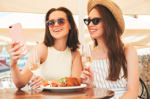 Dos jóvenes hermosas mujeres hipster sonrientes vestidas de verano Mujeres despreocupadas posando en el café de la terraza en la calle Modelos positivos bebiendo vino blanco con sombrero Mirando la pantalla del teléfono inteligente usando aplicaciones