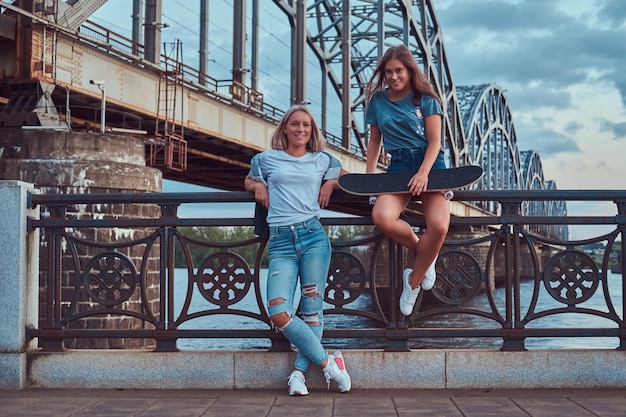 Dos jóvenes hipsters felices sostienen una patineta y se apoyan en una barandilla sobre el fondo del viejo puente.
