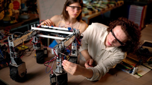 Dos jóvenes ingenieros arreglan un robot mecánico con ruedas en el taller con gafas industriales
