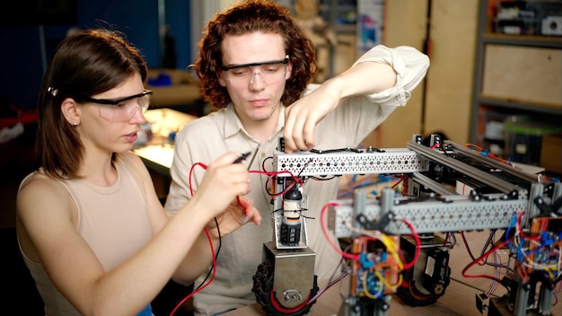 Dos jóvenes ingenieros arreglando un robot mecánico con ruedas en el taller con gafas industriales