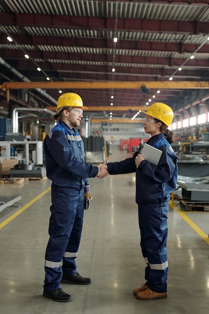 Dos jóvenes ingenieros exitosos en cascos protectores y ropa de trabajo mirando el uno al otro mientras se dan el apretón de manos dentro de un gran taller