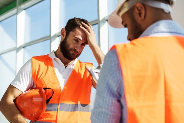 Dos jóvenes ingenieros masculinos en uniforme y cascos que trabajan en el sitio de construcción