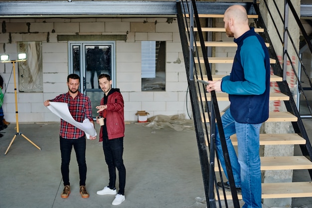 Dos jóvenes ingenieros con planos discutiendo los detalles del boceto mientras uno de ellos explica qué se puede cambiar sobre la construcción