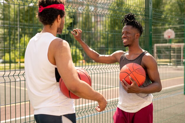 Dos jóvenes jugadores de baloncesto multiculturales interactuando después del juego mientras están de pie junto a la valla
