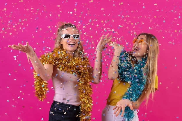Dos jóvenes mujeres caucásicas rubias de fiesta divirtiéndose lanzando confeti aislado en un fondo rosa