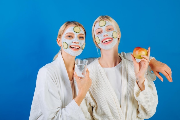 Dos jóvenes mujeres felices con máscara facial y rodajas de pepino en la cara van a un estilo de vida saludable ...