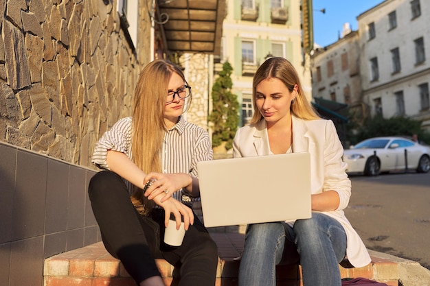 Dos jóvenes mujeres sonrientes mirando el monitor de la computadora portátil sentados al aire libre