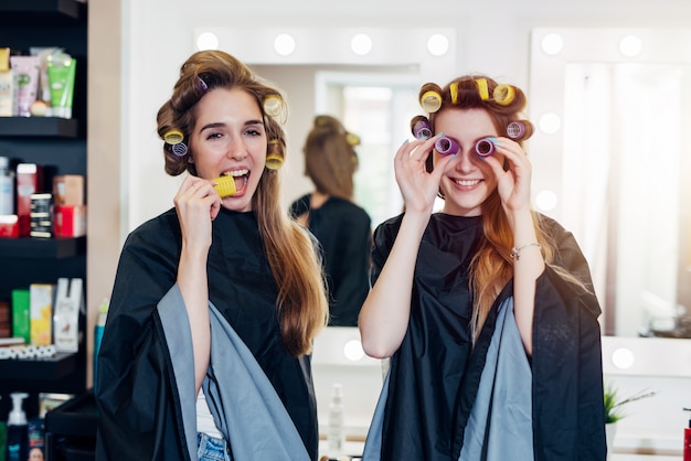 Dos jóvenes novias divertidas en Rizadores para el cabello con capas divirtiéndose juntos en el salón de belleza.