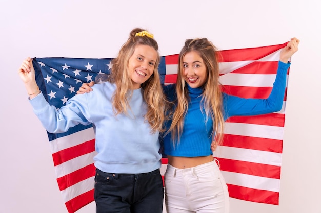 Dos jóvenes rubias caucásicas con bandera estadounidense sonriendo y divirtiéndose aisladas en un fondo blanco