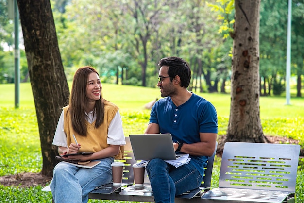 Foto dos jóvenes sentados en el parque charlando alegremente sosteniendo computadoras portátiles y bebiendo café