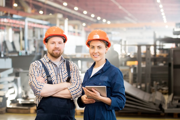 Dos jóvenes técnicos en ropa de trabajo y cascos protectores que trabajan en grupo en la fábrica.