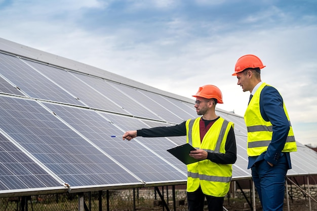 Dos jóvenes trabajadores con monos y cascos comprueban los paneles solares instalados Concepto de electricidad verde