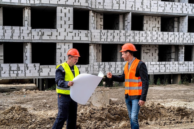 Dos jóvenes trabajadores uniformados en un sitio de construcción se paran con un plan y lo comparan con el nuevo edificio Concepto de construcción