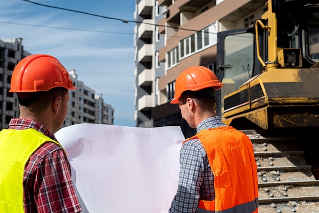 Foto dos jóvenes trabajadores en uniforme en un sitio de construcción se colocan con un plan y se comparan con el nuevo edificio. concepto de construcción
