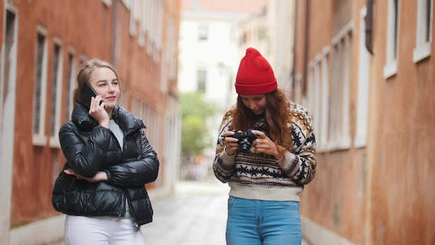 Dos jóvenes viajeras con ropa de abrigo caminando por las calles estrechas Una mujer hablando por teléfono y otra revisando sus tomas en la cámara