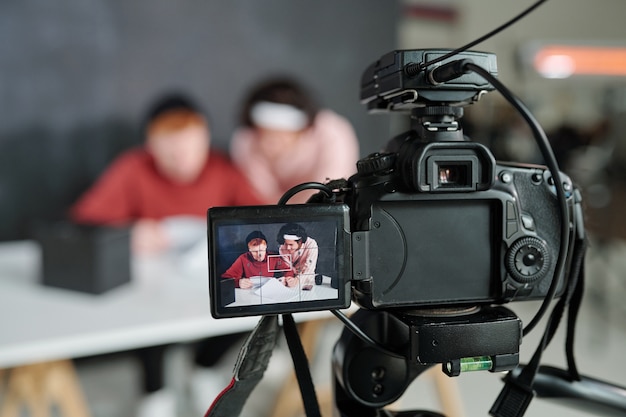 Dos jóvenes vloggers masculinos contemporáneos en la pantalla de la cámara de vídeo de pie delante del escritorio en el estudio