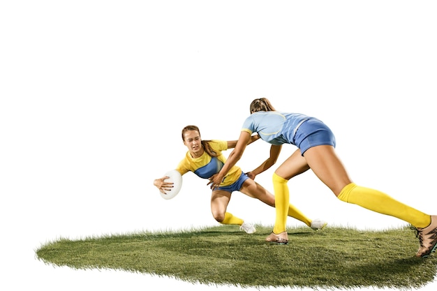Los dos jugadores de rugby femeninos jóvenes aislados sobre fondo blanco de estudio con hierba verde. Emociones humanas. Chicas caucásicas en forma. Competición deportiva, movimiento, movimiento, lucha, confrontación, ataque