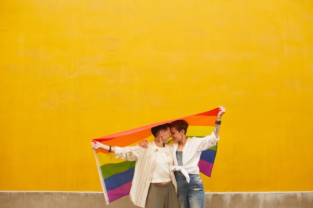 Foto dos lesbianas felices enamoradas que están de pie contra la pared amarilla al aire libre y sosteniendo la bandera de colores