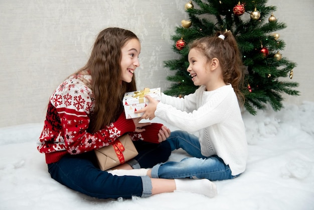 Dos lindas chicas sonrientes se sientan al lado del árbol de Navidad y se dan regalos de Año Nuevo Navidad