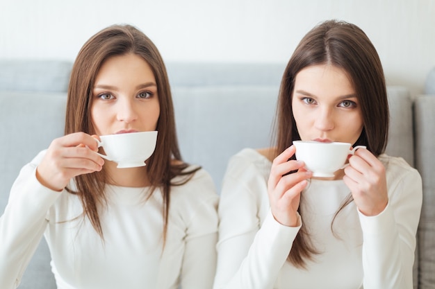 Dos lindas y encantadoras hermanas gemelas jóvenes sentados en el sofá y tomando café