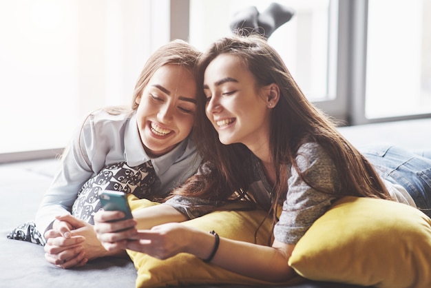 Dos lindas hermanas gemelas sonrientes sosteniendo smartphone y haciendo selfie. Las chicas se acuestan en el sofá posando y alegres