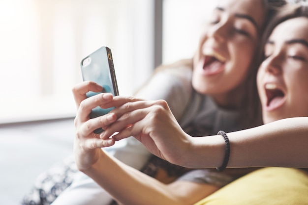 Dos lindas hermanas gemelas sonrientes sosteniendo smartphone y haciendo selfie. las mujeres se acuestan en el sofá posando y alegría