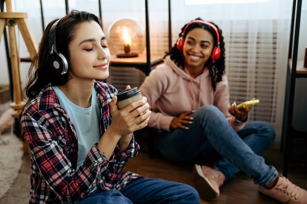 Dos lindas mujeres disfrutan escuchando música con café en casa. Bonitas novias en auriculares se relajan en la habitación, amantes del sonido descansando en el sofá, ocios de amigas juntos