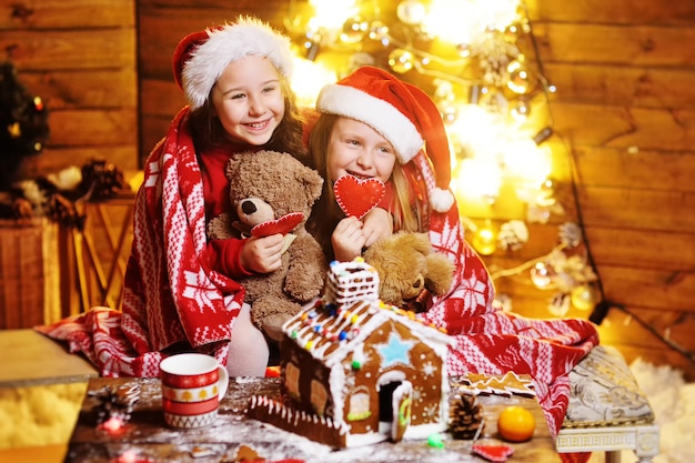 Dos lindas niñas divertidas con sombreros rojos de Papá Noel cubiertos con una sonrisa a cuadros, decoración navideña y luces y hacen una hermosa casa de pan de jengibre