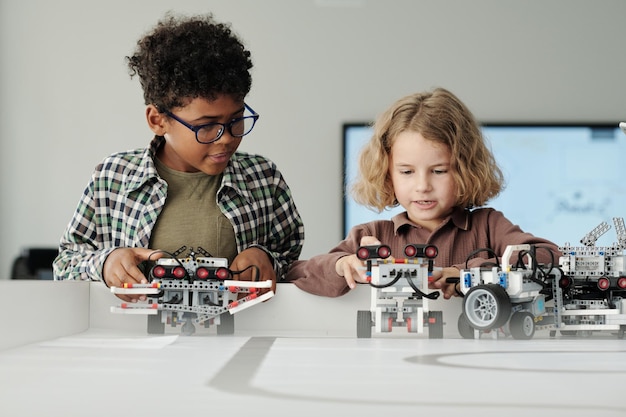 Dos lindos colegiales interculturales jugando con robots de juguete hechos a mano por mesa