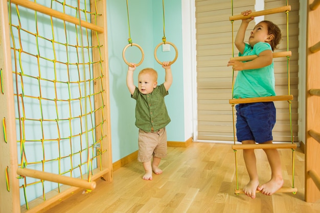 Dos lindos hermanos jugando en el gimnasio deportivo de madera en casa Chicos descalzos subiendo escaleras de madera y anillos Creciendo fuerte y saludable concepto