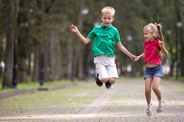 Dos lindos jóvenes sonrientes divertidos niños, niña y niño, hermano y hermana, saltando y divirtiéndose en borrosa brillante parque soleado.