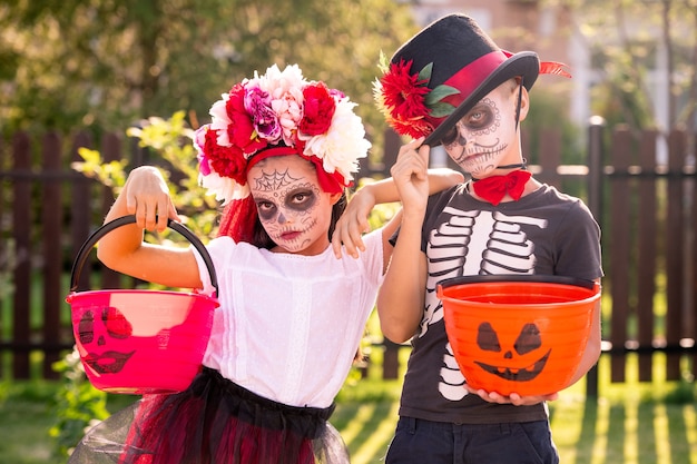 Dos lindos niños pequeños de halloween con caras pintadas sosteniendo cubos con dulces mientras están parados uno cerca del otro frente a la cámara