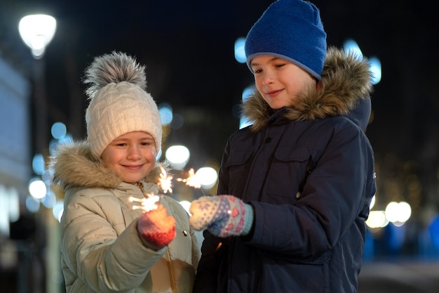 Dos lindos niños pequeños, niño y niña en ropa de invierno abrigada sosteniendo fuegos artificiales de bengala en la noche oscura al aire libre bokeh. Concepto de celebración de año nuevo y Navidad.