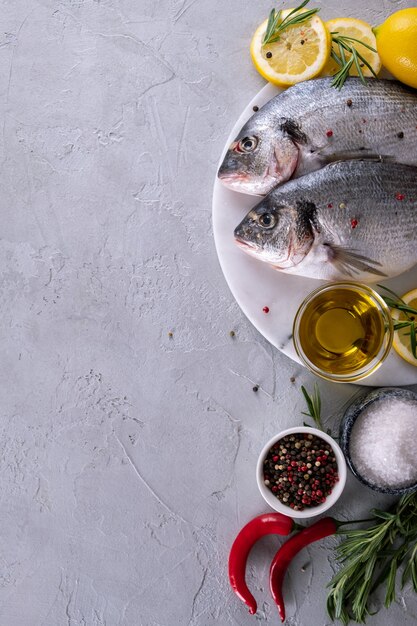 Dos listos para cocinar pescado crudo besugo con hierbas, limón y aceite de oliva en tablero de mármol sobre fondo gris. Con copia espacio. Vista superior