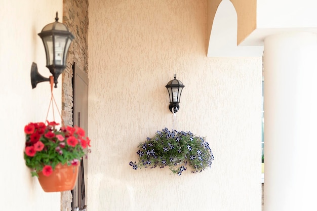 Dos macetas con flores rojas y azules en una pared de color amarillo pálido colgando de dos linternas junto a la puerta de la casa en un día soleado de verano