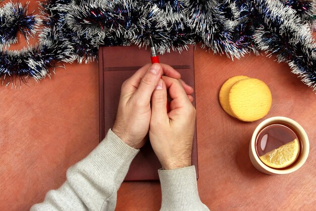 Las dos manos con bolígrafo rojo acostado sobre un cuaderno en el escritorio de oficina con té y galletas